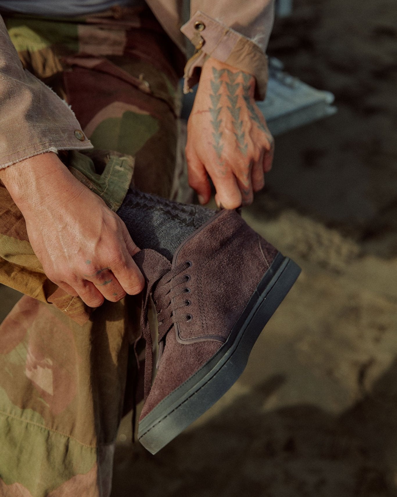 A close-up lifestyle shot of Jason Momoa wearing a purple suede lace-up ankle boot, pulling up their jeans, with a camouflage shirt and jacket visible in the background.