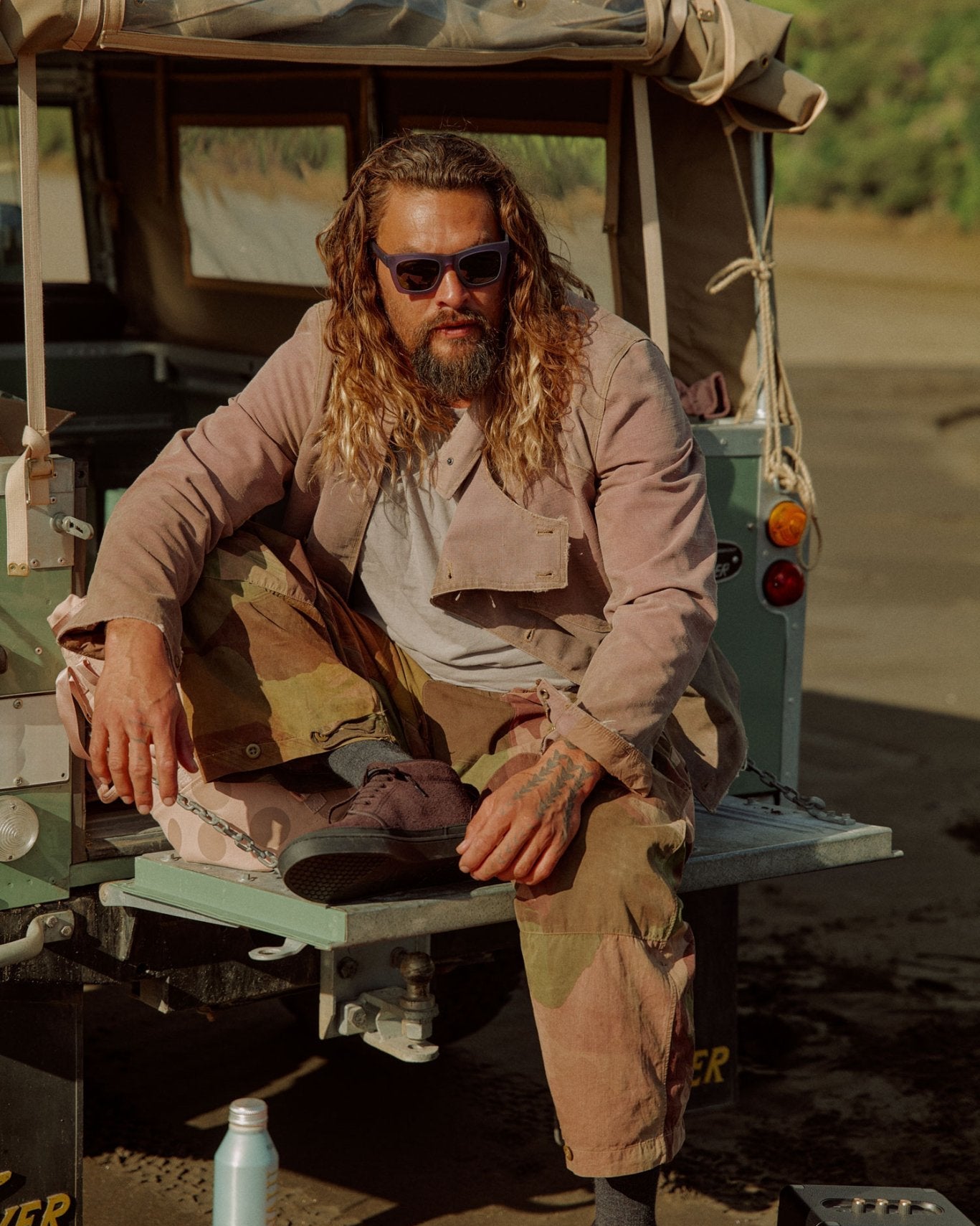 Jason Momoa, wearing sunglasses, a beige jacket, and camouflage pants sits on the tailgate of a vintage green vehicle outdoors.